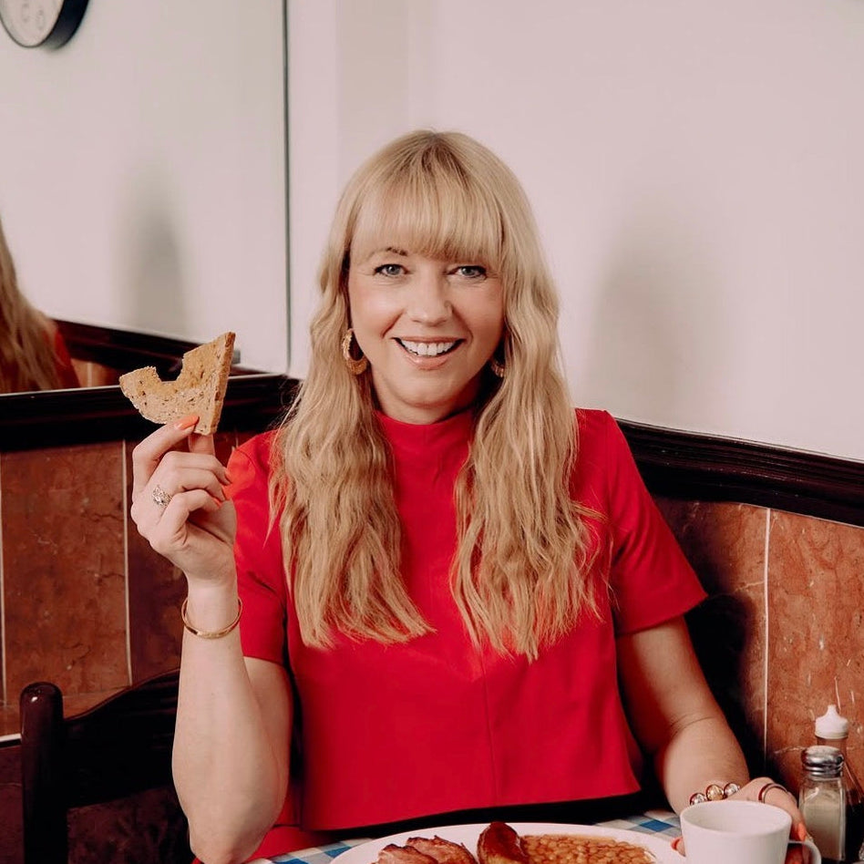A smiling SARA COX wearing LOEL Small Gold Tassel Hoop earrings at a breakfast table in a diner, holding a slice of toast. Plates with food, a mug, condiments, wall clocks, and a mirror are visible in the scene.