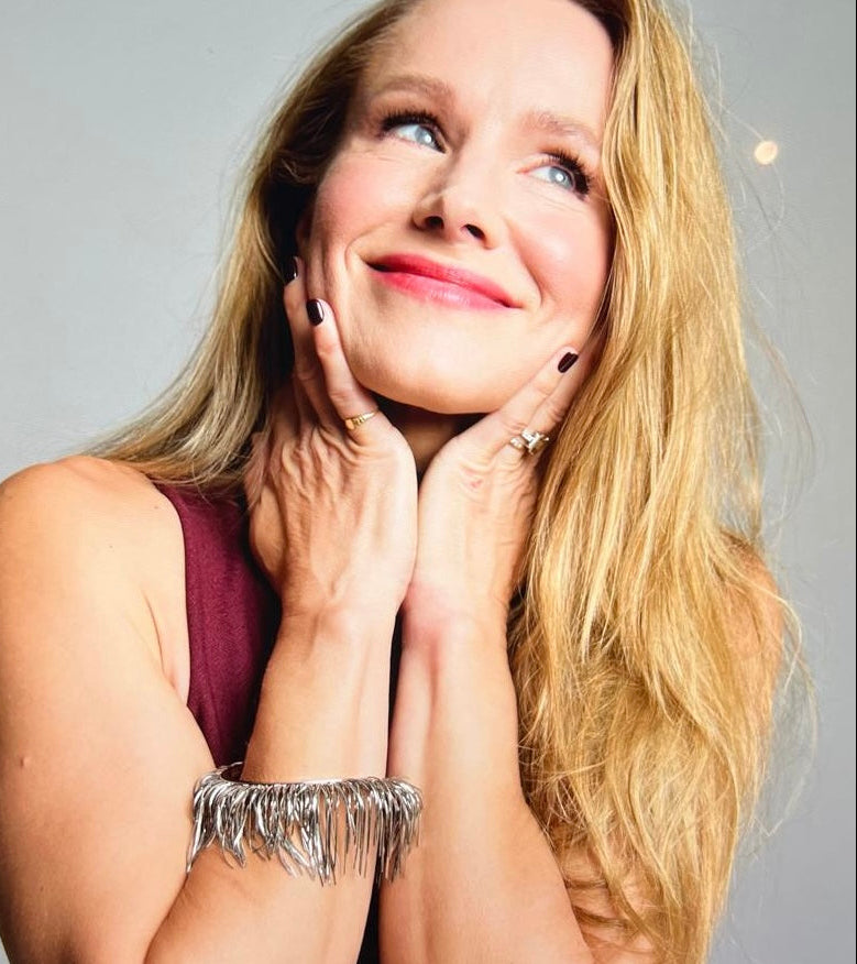 A close-up portrait of a smiling Kristen Bell wearing LOEL Feather cuff silver fringe bracelet, with her hands resting on her cheeks against a plain light grey background.